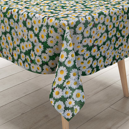 Rechteckige Wachstuch-Tischdecke mit dichtem Gänseblümchen-Muster. Zahlreiche weiße Blüten mit gelber Mitte bedecken einen grünen Hintergrund aus Blättern und Gräsern und erzeugen die Optik einer blühenden Sommerwiese. Zwischen den Blüten sind vereinzelt kleine Marienkäfer zu sehen, die dem floralen Design einen natürlichen und verspielten Akzent verleihen. Die Tischdecke fällt über die Kanten eines Tisches herab.