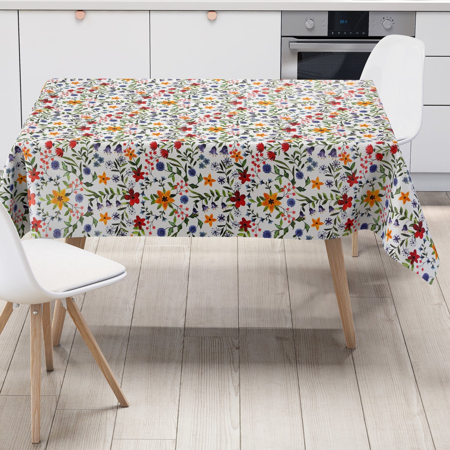 Weiße Wachstuch-Tischdecke mit dichtem, farbenfrohem Wildblumenmuster. Rote, gelbe, orangefarbene, blaue und violette Blüten sowie grüne Blätter und Ranken verteilen sich gleichmäßig über die gesamte Oberfläche und erinnern an eine blühende Sommerwiese im Landhausstil. Die rechteckige Tischdecke ist dekorativ über einen Tisch drapiert.