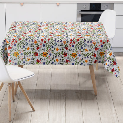 Weiße Wachstuch-Tischdecke mit dichtem, farbenfrohem Wildblumenmuster. Rote, gelbe, orangefarbene, blaue und violette Blüten sowie grüne Blätter und Ranken verteilen sich gleichmäßig über die gesamte Oberfläche und erinnern an eine blühende Sommerwiese im Landhausstil. Die rechteckige Tischdecke ist dekorativ über einen Tisch drapiert.