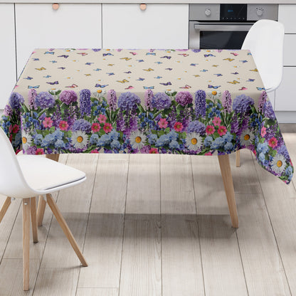 Wachstuch-Tischdecke mit cremefarbenem Hintergrund, dekoriert mit bunten Schmetterlingen und weißen Margeriten. Entlang der Ränder verläuft eine üppige Blumenbordüre aus Lavendel, Hortensien und weiteren Blüten in Violett-, Blau-, Rosa- und Weißtönen. Das frühlingshafte Design erinnert an eine blühende Gartenwiese und sorgt für eine freundliche, sommerliche Atmosphäre.