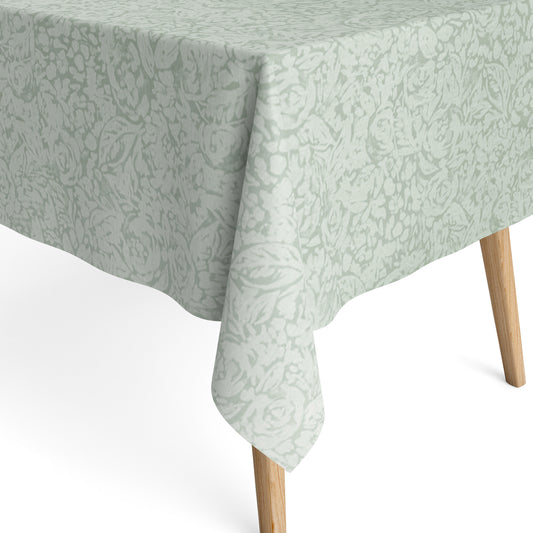Beschichtete Baumwoll-Tischdecke in hellem Salbeigrün mit floralem Allover-Muster aus Blüten und Blättern auf hellem Hintergrund, über eine Tischecke drapiert. Die matte Oberfläche wirkt pflegeleicht, die Tischbeine aus hellem Holz sind sichtbar.