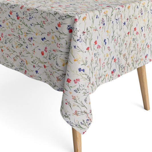 Beschichtete Baumwoll-Tischdecke mit zartem Streublumenmuster auf hellem Hintergrund. Filigrane Wildblumen und Gräser in Rot, Blau, Gelb und Rosa verteilen sich gleichmäßig über die Oberfläche. Über eine Tischecke drapiert, mit sichtbaren hellen Holzbeinen.