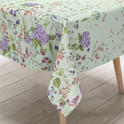 Wachstuchtischdecke mit romantischem Blumenmuster auf mintgrünem Hintergrund. Zarte Wildblumen, filigrane Zweige und Blütenranken in Rosa, Blau, Lila und Rot verteilen sich locker über die Fläche. Große violette Hortensien setzen dekorative Akzente entlang der oberen und unteren Bereiche. Das detailreiche, aquarellartige Dessin wirkt leicht, natürlich und verspielt und verleiht dem Tisch eine frische, florale Ausstrahlung.