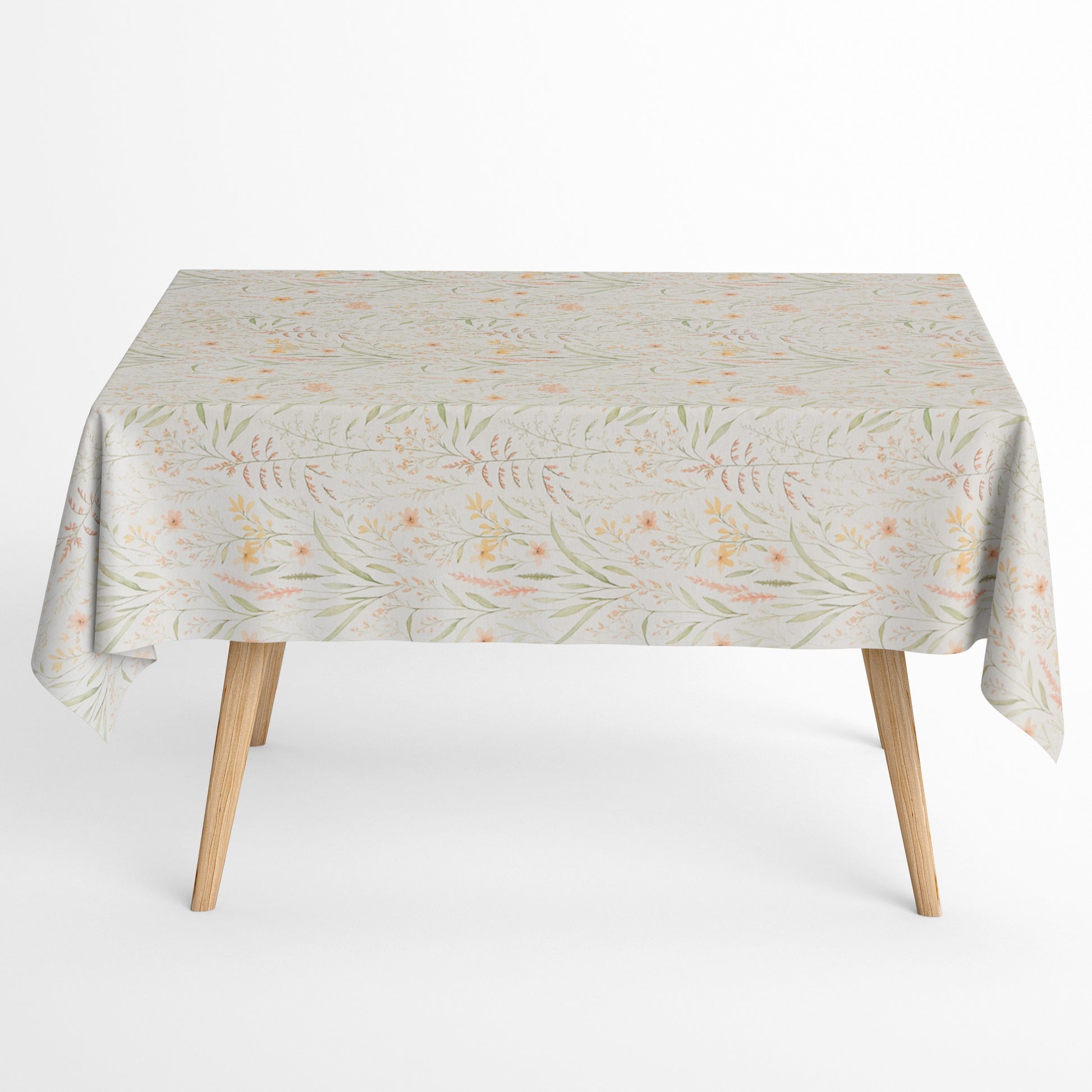 Beschichtete Baumwoll-Tischdecke mit filigranem Naturmuster auf hellem Hintergrund. Zarte Gräser, Blätter und kleine Blüten in Apricot-, Gelb- und Grüntönen verteilen sich gleichmäßig über die Oberfläche. Über eine Tischecke drapiert, mit sichtbaren hellen Holzbeinen.
