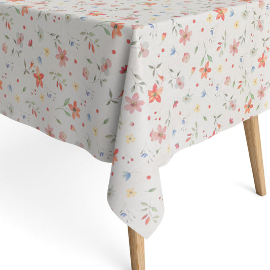Beschichtete Baumwoll-Tischdecke mit zartem Streublumenmuster auf hellem Hintergrund. Kleine Blüten, feine Zweige und dekorative Punkte in Rosa, Korall, Gelb, Blau und Grün verteilen sich gleichmäßig über die Oberfläche. Über eine Tischecke drapiert, mit sichtbaren hellen Holzbeinen.