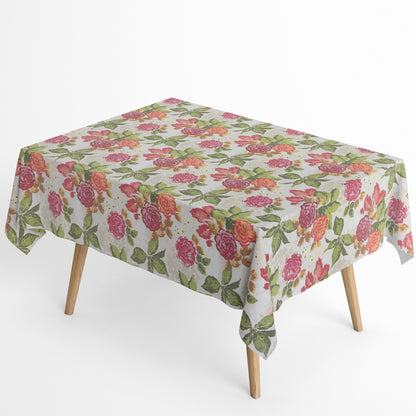 Beschichtete Baumwoll-Tischdecke mit großem floralem Muster auf hellem Hintergrund. Auffällige Blüten in Pink, Rot und Orange sowie grüne Blätter verteilen sich über die Oberfläche. Über eine Tischecke drapiert, mit sichtbaren hellen Holzbeinen.